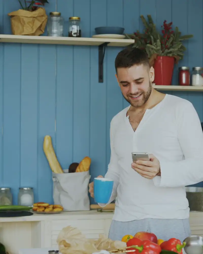 A man smiling at his phone while holding a coffee in a bright kitchen.